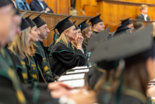 Kilka osób siedzi w drewnianej sali audytoryjnej podczas uroczystości. Wszyscy mają na sobie czarne togi z zielonymi lamówkami oraz czarne birety z chwostami. Na stołach przed nimi leżą dokumenty i długopisy. W tle widoczne są drewniane ławy oraz osoby siedzące na wyższych rzędach.