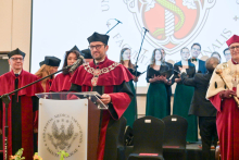 The dean, wearing a burgundy gown with a decorative chain, speaks from behind a lectern bearing the WUM coat of arms. Next to him stand people in burgundy gowns, and in the background a group of people in green dresses hold open songbooks