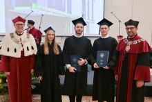 Five people. Two people in the middle are wearing black gowns and birettas, holding navy blue briefcases with the WUM coat of arms. On either side are two people wearing burgundy gowns.