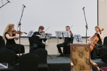 Musical performance. Musicians are seated on chairs, holding instruments such as violins in their hands. They are elegantly dressed.