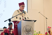 The rector, wearing a burgundy gown with a white collar, speaks from behind a lectern bearing the WUM coat of arms. Next to him are people in burgundy gowns, and at the bottom is a fragment of a bouquet of yellow flowers.