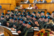 Grupa osób w czarnych togach z zielonymi lamówkami i czarnych biretach z chwostami siedzi w drewnianej sali audytoryjnej podczas uroczystości. Na stołach przed nimi leżą dokumenty i długopisy. Część osób klaszcze, a w tle widoczne są kolejne rzędy z drewnianymi ławami, na których siedzą inni uczestnicy. lomy