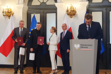 Pięć osób stoi w eleganckiej sali przed flagami Polski i Unii Europejskiej, dwie osoby trzymają bordowe teczki i białe torby. Po prawej stronie znajduje się mównica z napisem „Ministerstwo Zdrowia”.