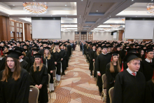 Hundreds of people in black gowns and caps sit on chairs arranged in long rows on both sides of a wide aisle.