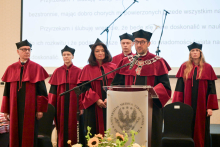 Six people in burgundy gowns and black caps standing on stage. In the foreground. Behind the lectern stands the dean dressed in a burgundy gown. 