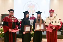 Four people pose for a photo. The two people in the middle are wearing black gowns and caps, holding diplomas in brown frames. On either side are two men—the dean and the rector. They are holding red folders in their hands.