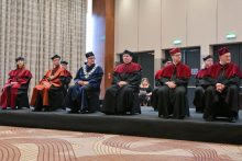 Seven people in long robes sit on a raised stage. 