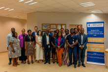 Grupa osób stoi w pomieszczeniu biurowym obok roll-upu z napisem „University of Ghana Medical Centre Ltd” i informacją o wsparciu dla jednostki badań klinicznych. 