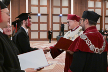The moment of awarding the diploma. A young man in a black gown and cap shakes hands with the rector in a burgundy gown. Next to him stands the dean in a burgundy gown