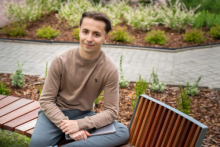 Adrian Bednarek na Kampusie Banacha. Siedzi na drewnianej ławce w ogrodzie z roślinami ozdobnymi. Osoba ma na sobie beżowy sweter i szare spodnie, trzyma zamknięty laptop na kolanach. W tle widać ścieżkę z kostki i rabaty z zielonymi krzewami.