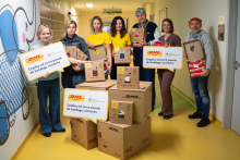 W jasnym szpitalnym korytarzu stoi grupa osób z dużymi kartonowymi pudełkami i tablicami. Na pudełkach widnieją kolorowe grafiki przedstawiające wełniane czapki. 