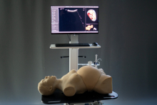 Na pierwszym planie znajduje się realistyczny fantom medyczny w pozycji leżącej na czarnym stole. Za nim stoi monitor na białym stojaku, na którym wyświetlony jest obraz ultrasonograficzny – widoczny jest przekrój anatomiczny oraz kolorowa wizualizacja płodu w macicy. 