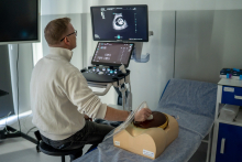 Osoba siedzi na stołku przed aparatem ultrasonograficznym z dwoma ekranami, na których widoczny jest obraz USG w odcieniach szarości przedstawiający przekrój anatomiczny. Na stole obok znajduje się realistyczny fantom z przezroczystą pokrywą, w którym wykonywana jest symulacja badania ultrasonograficznego przy użyciu głowicy.