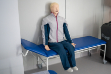 Fantom medyczny w realistycznym ubraniu – szarej kurtce z fioletową lamówką i granatowych spodniach – siedzi na niebieskim leżance z metalową ramą i kółkami. 