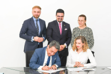 Na zdjęciu pięć osób znajduje się w jasnym pomieszczeniu przy szklanym stole. Na pierwszym planie jedna osoba w granatowym garniturze podpisuje dokumenty, a obok siedzi osoba w białej koszuli, patrząca na dokumenty. 