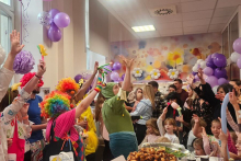 Grupa osób, dorosłych i dzieci, ubranych na kolorowo, świętuje w dużej jasnej sali. Wokół nich są fioletowe baloniki, na ścianach widać rysunki.