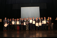 Grupa elegancko ubranych osób. Stoją na scenie i pozują do zdjęcia. Część z nich trzyma w rękach dyplomy oprawione w ramki.