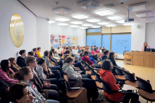 Jedna osoba stoi przy mównicy w sali i prowadzi prezentację. W sali siedzi kilkadziesiąt osób, które patrzą w stronę ekranu i mównicy. Na ekranie widoczny jest slajd z tytułem „Sfałszowane produkty lecznicze – skład i niebezpieczeństwo dla zdrowia i życia” oraz schemat z napisami.