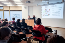 Dwie osoby stoją przy mównicy i prowadzą prezentację. Jedna osoba stoi przy komputerze, druga wskazuje na ekran z wyświetlonym slajdem. Na slajdzie widoczny jest tytuł „Sfałszowane produkty lecznicze – skład i niebezpieczeństwo dla zdrowia i życia” oraz schemat z napisami. W sali siedzi grupa osób.