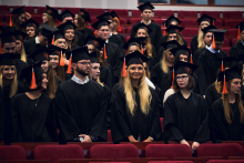 A group of people dressed in black academic gowns and caps with orange tassels stand in rows in the auditorium. 