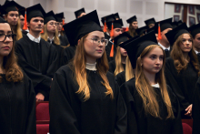 People dressed in black academic gowns and caps with orange tassels stand in rows in the auditorium.