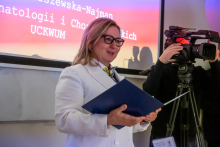 Kobieta w białym fartuchu lekarskim stoi i przemawia. W rękach trzyma rozłożoną teczkę. W tle widać człowieka z kamerą. Za nimi ekran multimedialny.