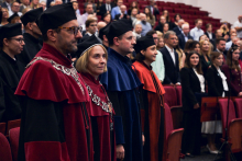People wearing burgundy, navy blue, and orange academic gowns stand in the front row of the auditorium. Decorative silver ceremonial chains are visible on the gowns.