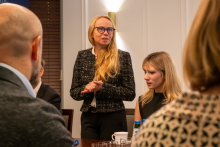 Osoba w eleganckim żakiecie stoi przy stole w sali konferencyjnej, trzyma dłonie złożone przed sobą.