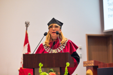 A person wearing a burgundy academic gown and cap stands behind a lectern with a microphone. A decorative silver ceremonial chain is visible around their neck.