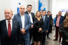 Grupa osób stoi w korytarzu przed drzwiami oznaczonymi tabliczką „Pracownia Tomografii Komputerowej”. Większość osób ubrana jest w eleganckie stroje.