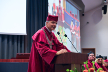 A person wearing a burgundy academic gown and cap stands behind a lectern with two microphones. A decorative silver ceremonial chain is visible around their neck.