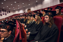 People dressed in black academic gowns and caps with orange tassels sit in rows of red armchairs in the auditorium. The caps feature round emblems with a symbol and inscription.