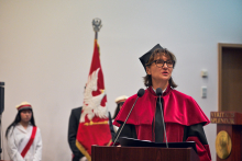 A person wearing a burgundy academic gown and cap stands behind a lectern with two microphones.