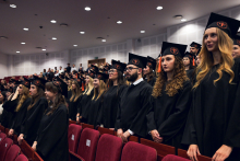 People dressed in black academic gowns and caps with orange tassels stand in rows of red chairs in the auditorium. The caps feature round emblems with a symbol and inscription. 