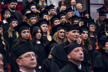 People dressed in black academic gowns and caps with orange tassels sit in rows of red armchairs in the auditorium. The caps feature round emblems with a symbol and inscription.