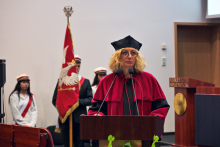 A person wearing a burgundy academic gown and cap stands behind a lectern with two microphones. 