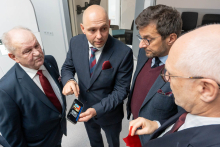 Kilka osób w eleganckich garniturach stoi w grupie w jasnym pomieszczeniu. Jedna z osób trzyma w dłoni smartfon, na którego ekranie widoczny jest kolorowy obraz medyczny - skan z tomografii komputerowej.