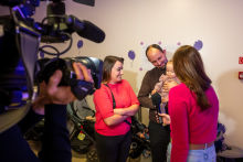 Cztery osoby stoją w pomieszczeniu przy jasnej ścianie z fioletowymi balonami na dekoracji. Jedna osoba trzyma małe dziecko na rękach. Osoba w czerwonym swetrze rozmawia z reporterką w czerwonym swetrze i jasnych spodniach, która trzyma mikrofon. Po lewej stronie widać kamerę i operatora. W tle stoją dwa wózki dziecięce.