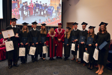 A group of people in black academic gowns and caps with orange tassels stand in a row, holding diplomas in navy blue folders and white printed bags.  