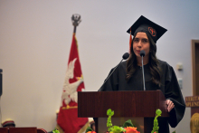 A person wearing a black academic gown and a cap with an orange tassel stands behind a wooden lectern with two microphones. 