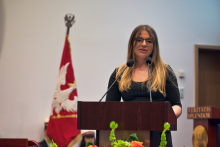 A person stands behind a wooden lectern with two microphones, wearing a black dress with short sleeves. 