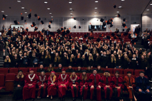 A large group of people in black gowns stand in the auditorium and throw their caps with orange elements into the air. In the front row, there are a dozen or so people in red and navy blue gowns with decorative chains; they are representatives of the university authorities.
