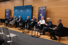 A discussion panel in a conference hall with eight participants seated in black armchairs arranged in a row. In front of each armchair, there is a small table with water bottles and glasses. In the background, a blue backdrop is visible with inscriptions highlighting the anniversaries.