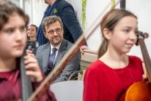 In the foreground, there are string instruments — a violin and a cello — held by children preparing for a performance. In the center of the frame, a man in an elegant suit with a tie is seated.