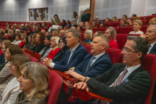 Several rows of people are seated in the hall on red chairs, looking toward the stage. Dozens of participants are attentively watching the event.