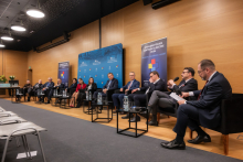 A discussion panel in a conference hall with eleven participants seated in black armchairs arranged along a wooden wall. In front of each armchair, there are small tables with water bottles and glasses. In the background, a blue backdrop is visible with inscriptions highlighting the anniversaries.