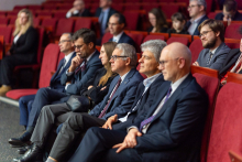 Several people are sitting on red armchairs in an auditorium, looking straight ahead