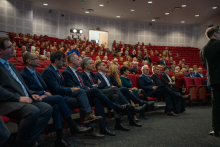 Several rows of people are seated in the hall on red chairs, looking toward the stage. Dozens of participants are attentively watching the event.