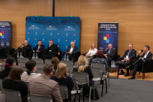 A discussion panel in a conference hall with eight participants seated in black armchairs arranged in a row. In front of each armchair, there are small tables with water bottles and glasses. In the background, a blue backdrop is visible with inscriptions highlighting the anniversaries.