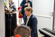 A man in an elegant blazer is playing the piano during an intimate concert. Next to him is an electronic keyboard, and in the background, observers can be seen, including a woman recording with her phone and a child in a red dress holding a violin.